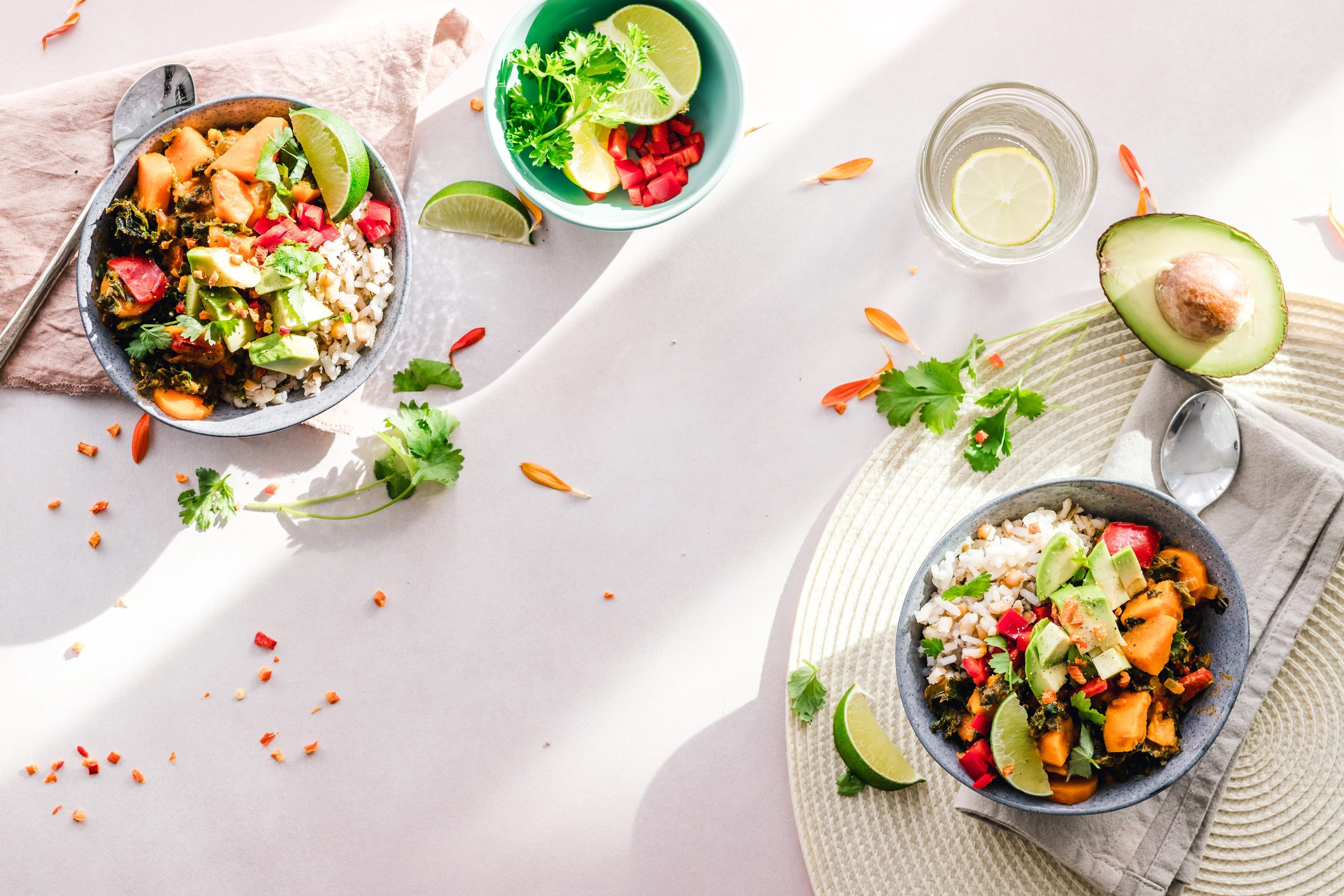 gesunde Ernährung Zeigt gesunde Nahrung für die Ernährungsberatung. Drei Schalen mit gesundem Essen auf einem Tisch mit weißer Tischdecke, eine Avocado und frische Kräuter und Blütenblätter, ein Glas frisches Wasser